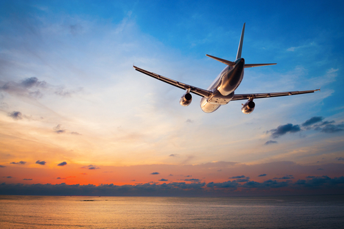 Airplane flying over water towards a colorful sunset sky.