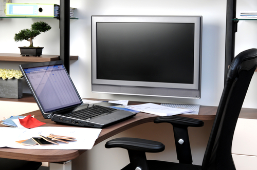 A cluttered desk with an open laptop, a wall-mounted monitor, and various items including papers, phones, and a bonsai tree.